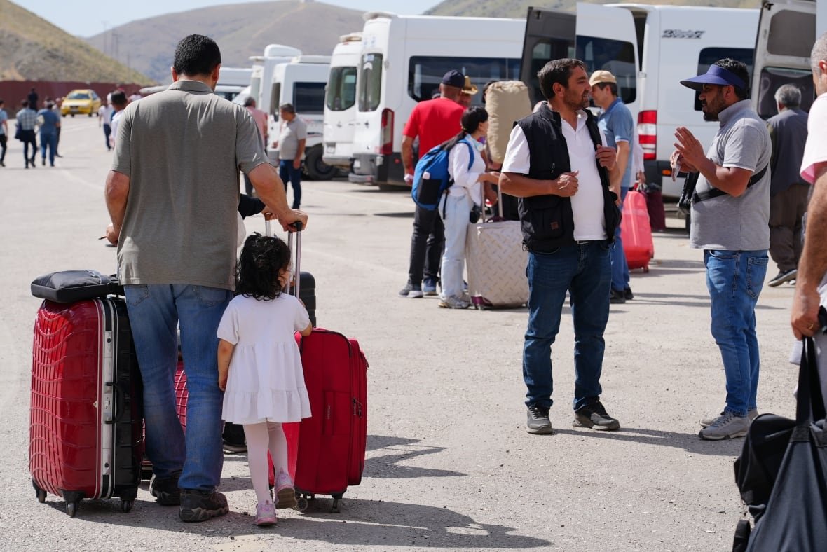 A man pushing red suitcases is seen walking with a young girl toward a row of white vans. 