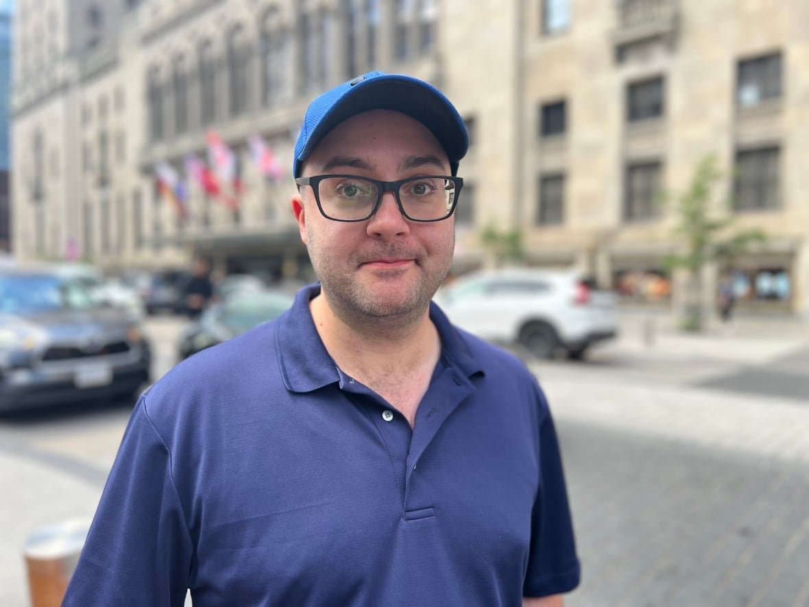 A man in a blue shirt with a blue ball cap and black glasses stands in front of a building.