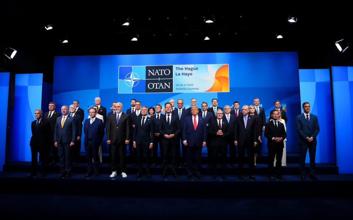 A group of world leaders wearing suits pose for a group photo in front of a screen that reads "NATO."