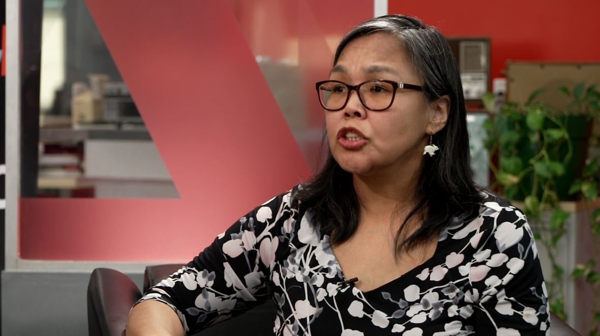 A woman with black, greying hair wearing glasses and a black shirt with a white flower pattern sitting in a chair