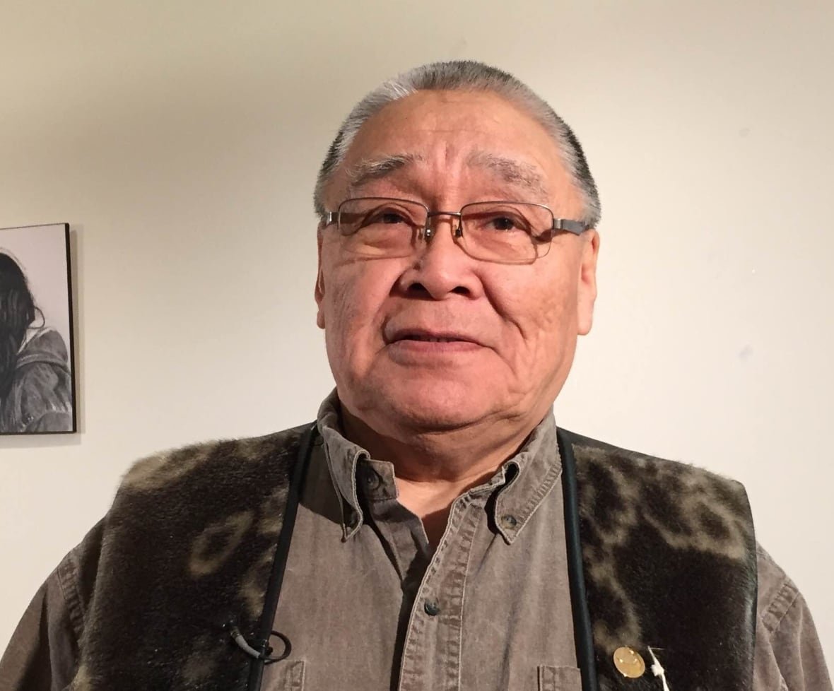 An older man in a sealskin vest stands in front of a wall with old photographs.