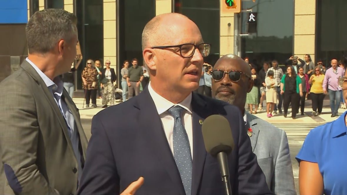A man in a suit speaks outside at a podium. A street corner is in the background.