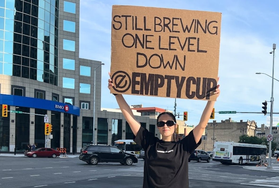 A woman stands at a street corner holding a cardboard sign above her head.