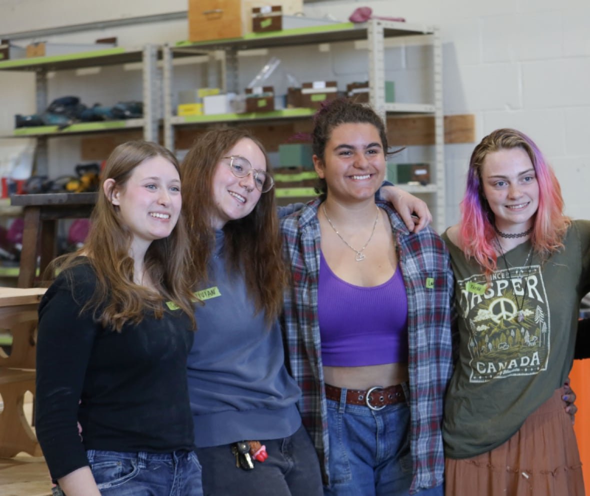 Four young women link arms for a photo in a woodworking shop.