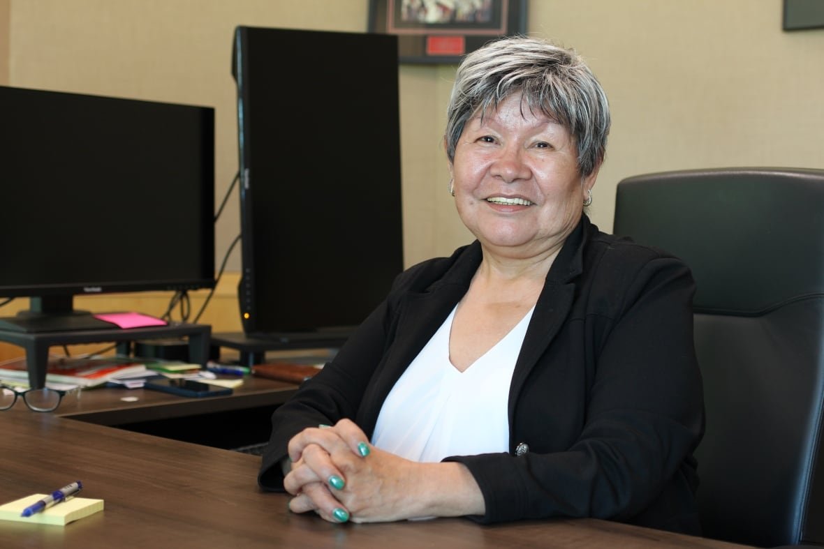A woman in a black blazer and white blouse poses for a photo while seated at her desk.