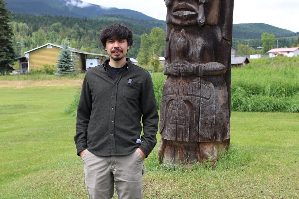 A man wearing a dark green button-down shirt and light green pants poses for a photo in front of a totem pole.