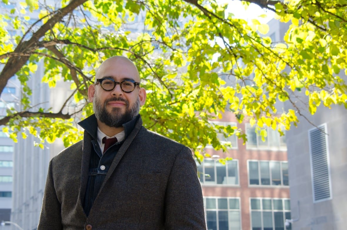 A bald, bearded man wearing a coat and tie standing in front of a tree and building.