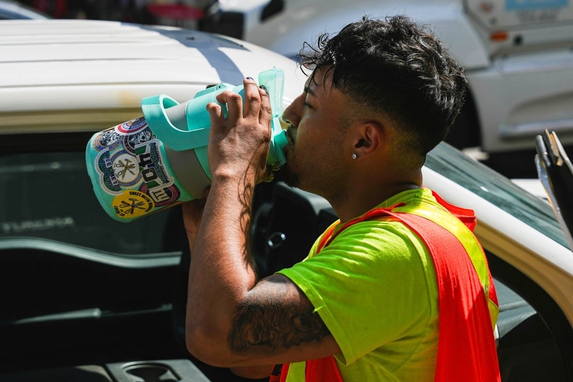 Wearing a yellow T-shirt and safety vest, a worker drinks from a giant canister near a vehicle, amid a heat wave.