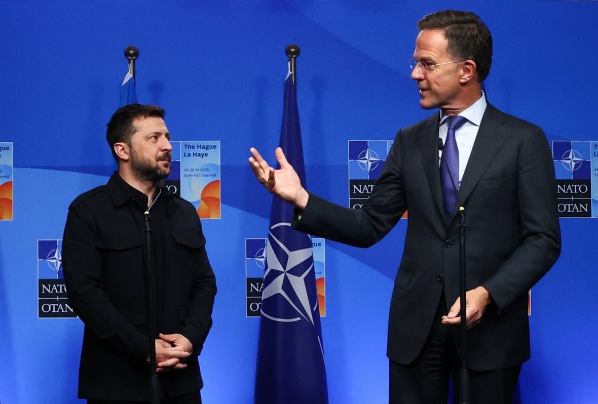 A man in a jacket with a beard stands next to a man in a suit near a NATO flag.