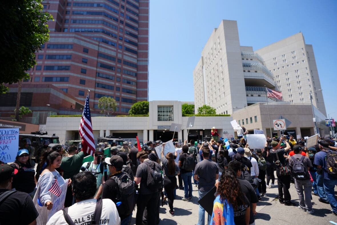 A crowd of people carrying American flags and protest signs gather outside a large building. 