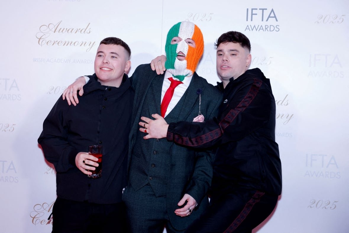 Three young men are shown hugging each other in a red carpet photo. One of them is wearing a ski mask over their head.