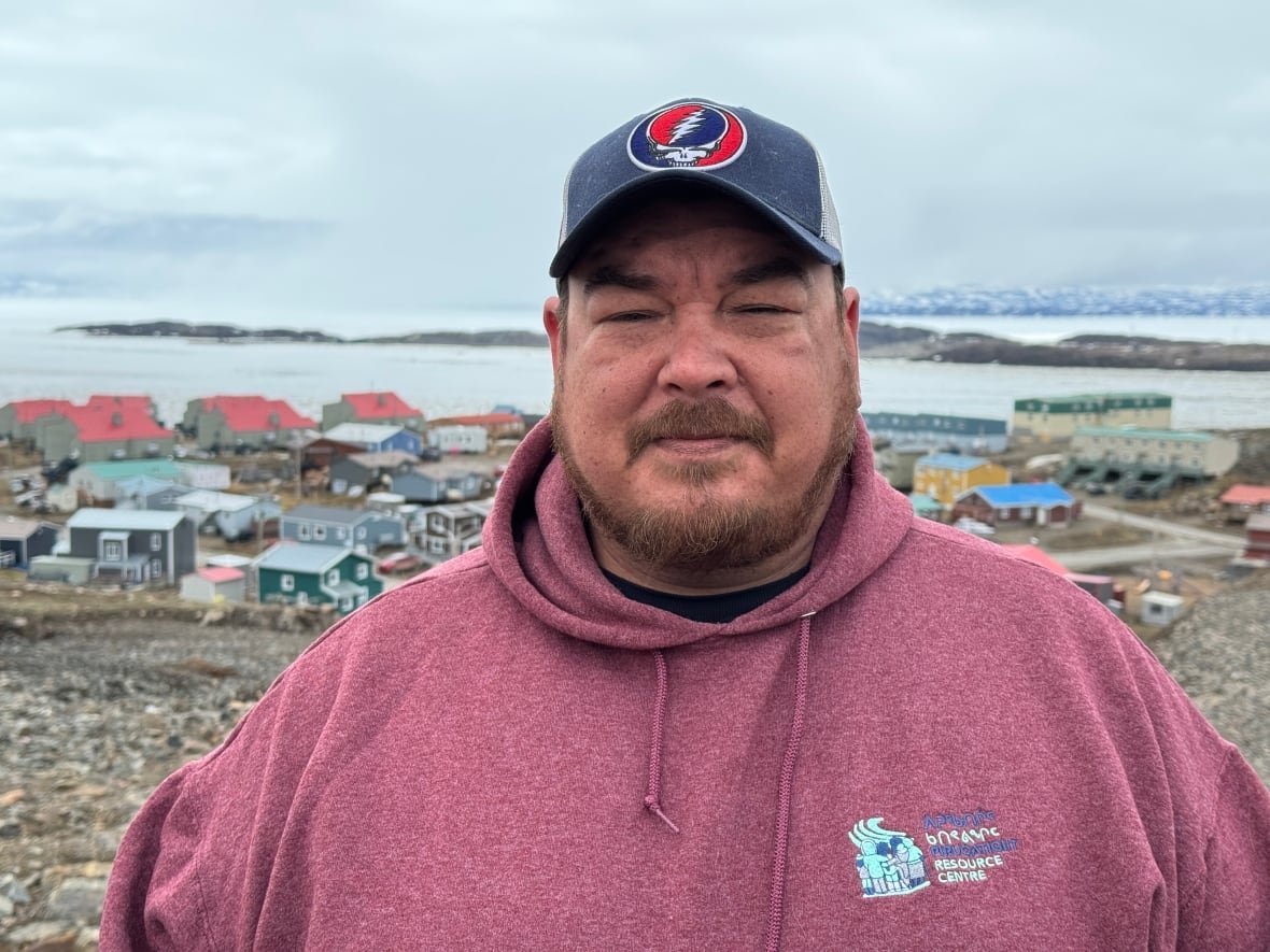A man in a hat and hoodie stands outside with an Arctic community in the background.
