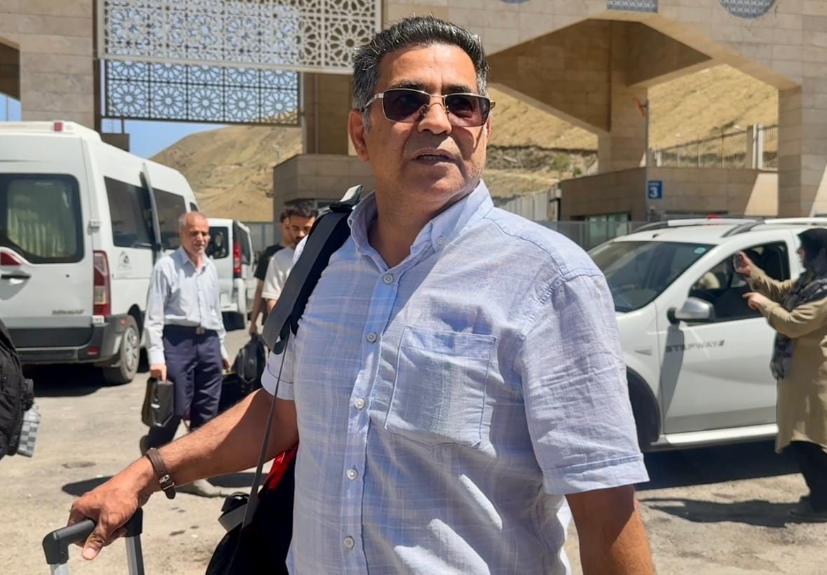 A man wearing a short-sleeve, blue checked shirt and sunglasses waits outside, his hand on a suitcase. Behind him are vans and other travellers at a border crossing.