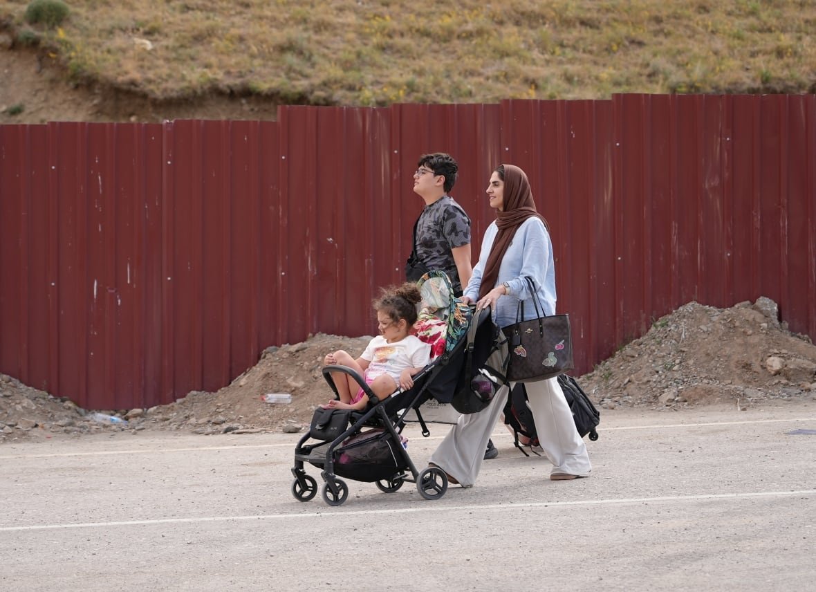 A woman wearing a brown headscarf pushes a black stroller, with a young child in it. along a road next to a red metal fence. Next to her walks a teenage boy, pulling a suitcase.