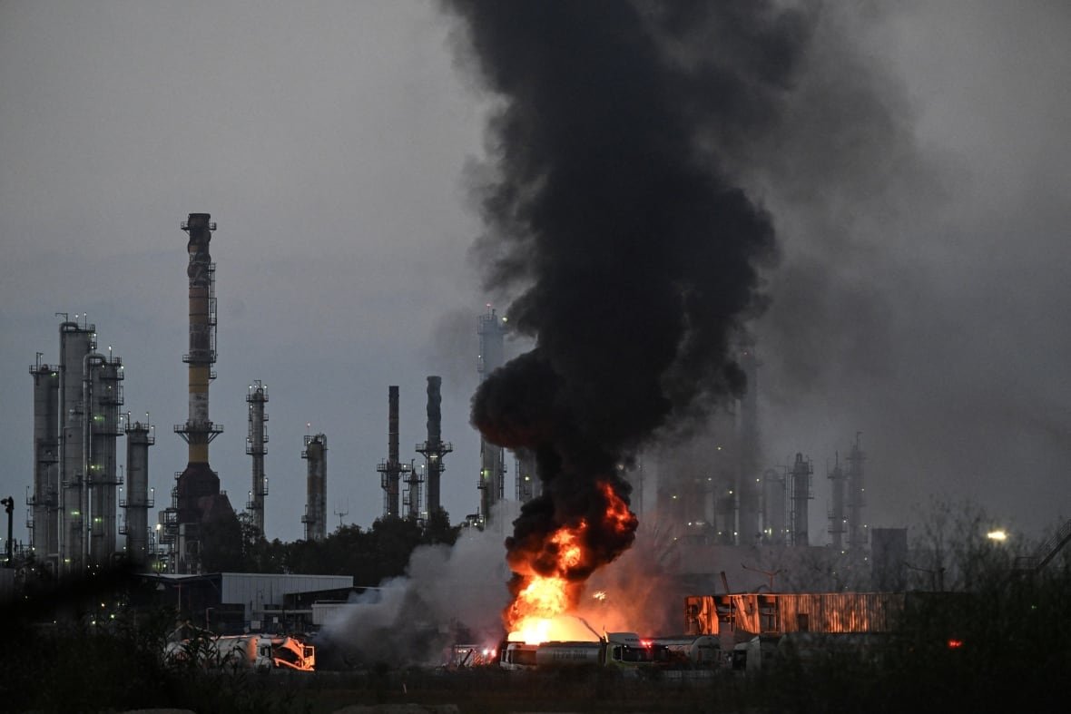 Black smoke rises from a fire in front of an industrial plant with many smokestacks.