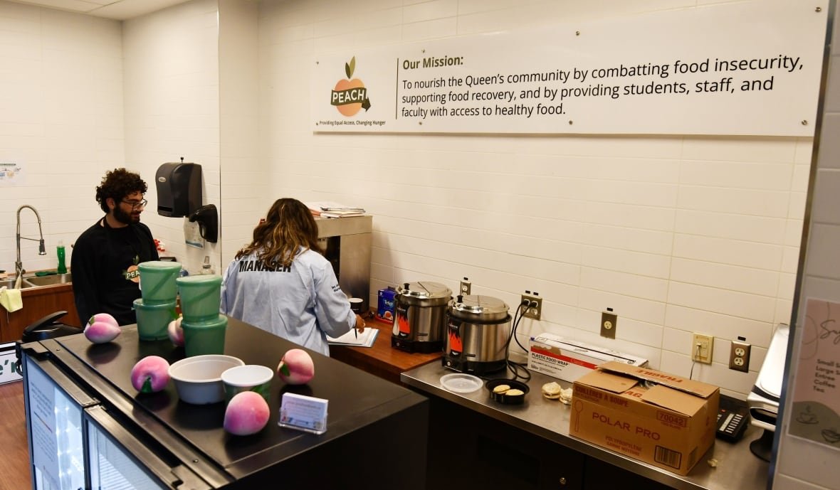 Two people are seen in what looks like a kitchen or the back of a cafe, with a large kitchen island with an inset mini-fridge, a sink, and cooking devices plugged in on a counter along the wall. A sign on the wall above declares that PEACH's mission is "To nourish the Queen's community"