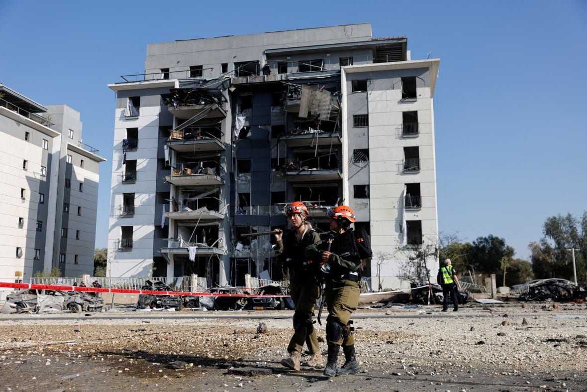 Two people wearing orange helmets stand in front of a damaged residential building. 