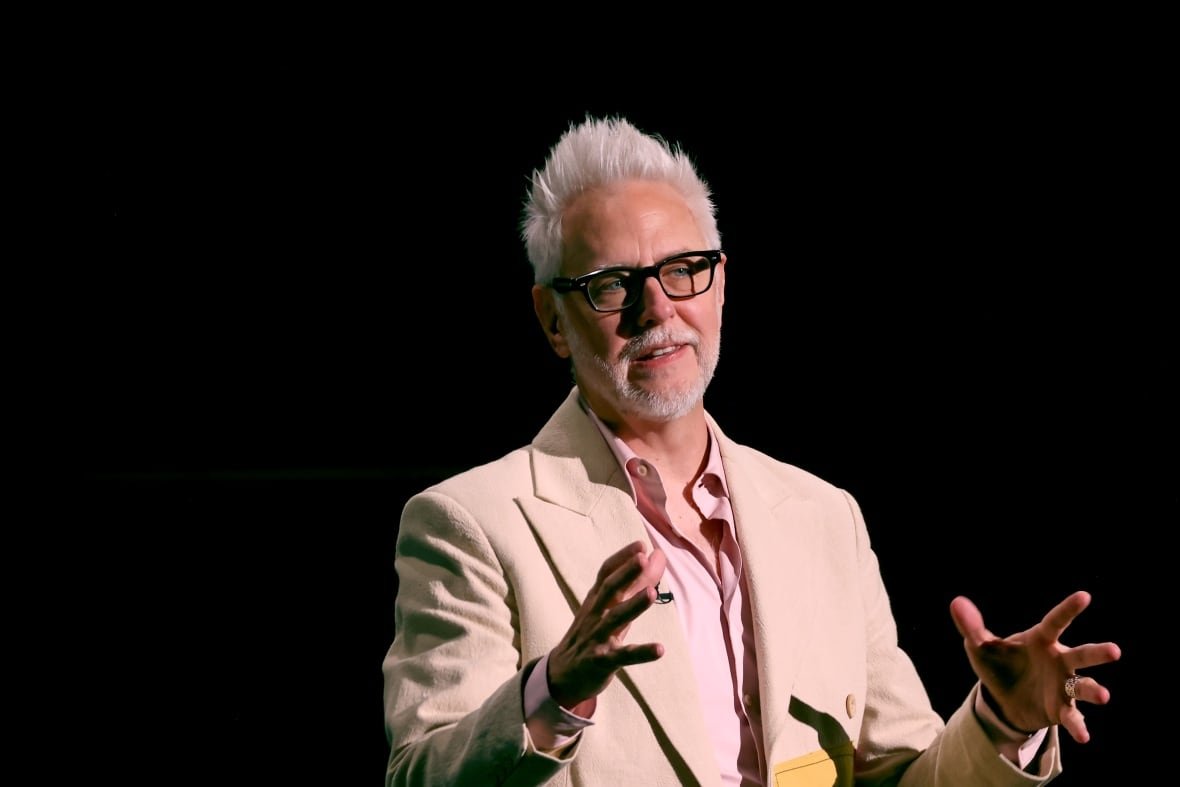 A man with white spiked hair, in a cream suit, gestures while speaking. 