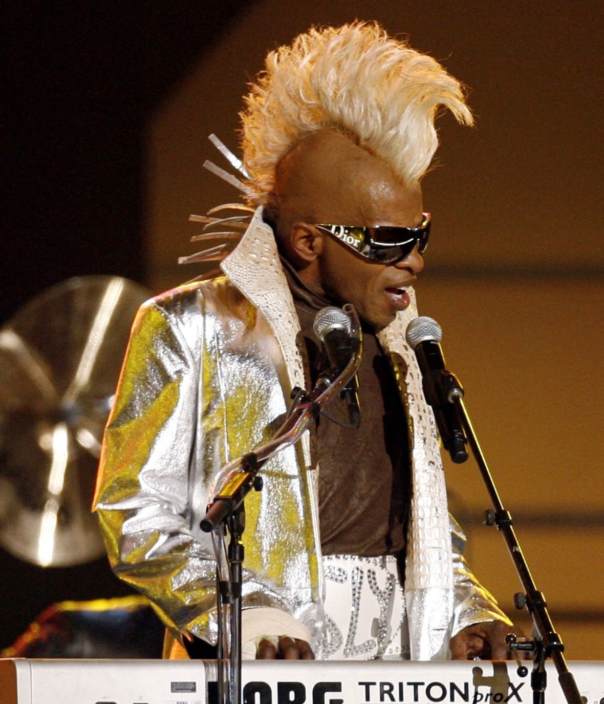 A man with a dramatic mohawk plays a keyboard and sings into a microphone. 