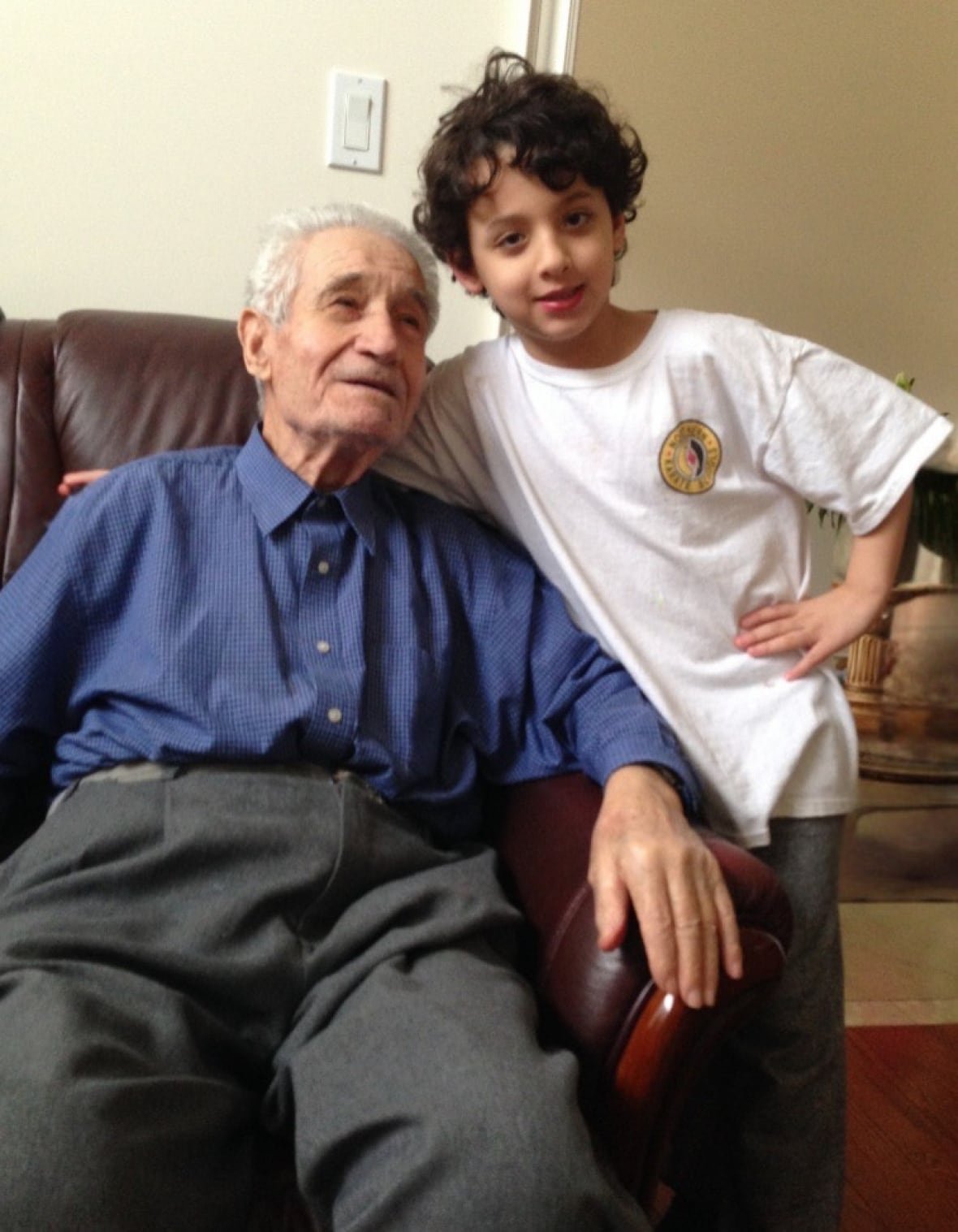 An elderly man sits in a red leather couch. He is wearing a blue button up shirt and grey slacks. He smiles up at a little boy standing next to the couch, arms around the elderly man. The boy is wearing a white t-shirt with jeans and smiling at the camera. 