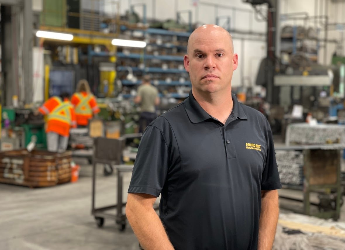 A bald man in a black polo poses for a photo in a factory floor setting as people in flourescent safety gear work behind him. 