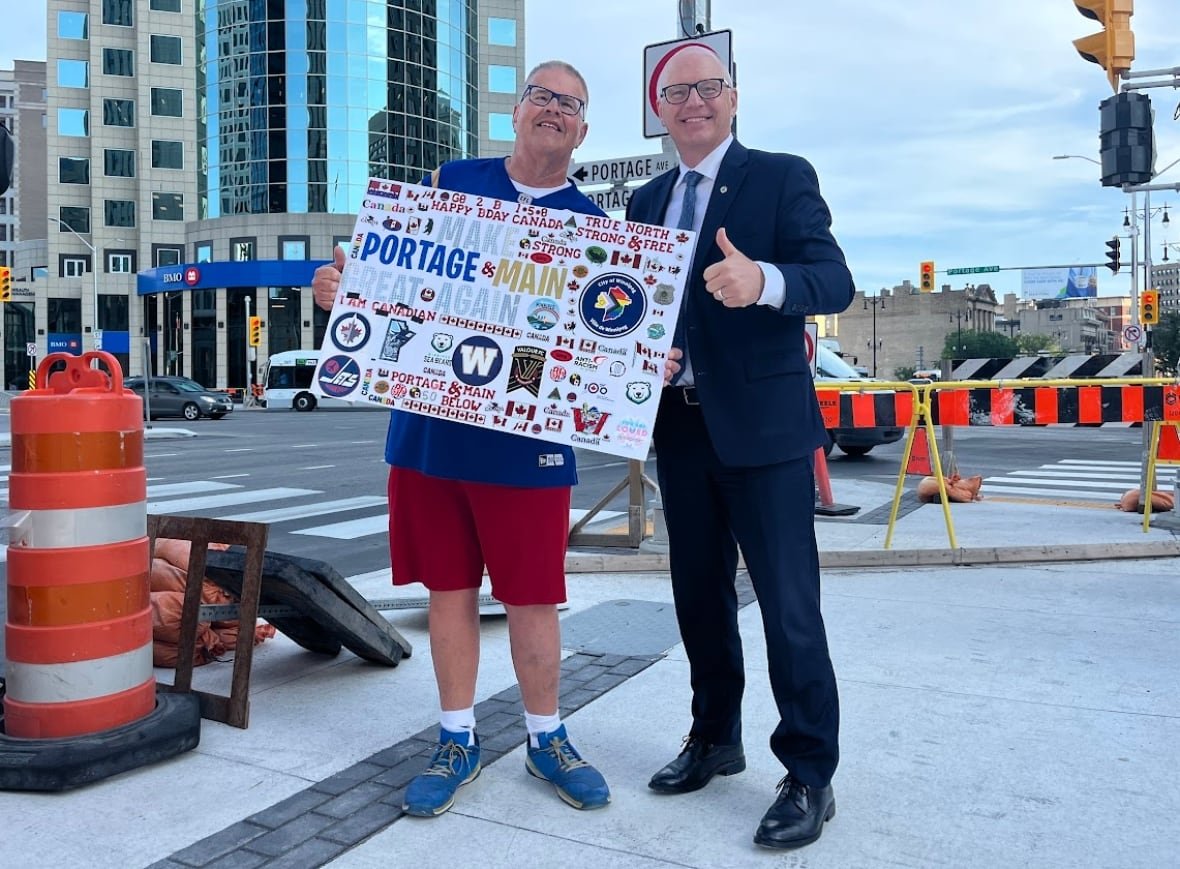 Two men, one in a suit and one wearing a sports jersey, pose and give the thumbs up sign