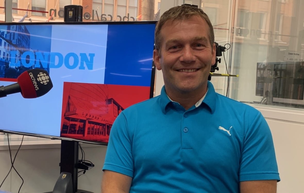 A man wearing a blue shirt sits with a TV screen behind his left side.