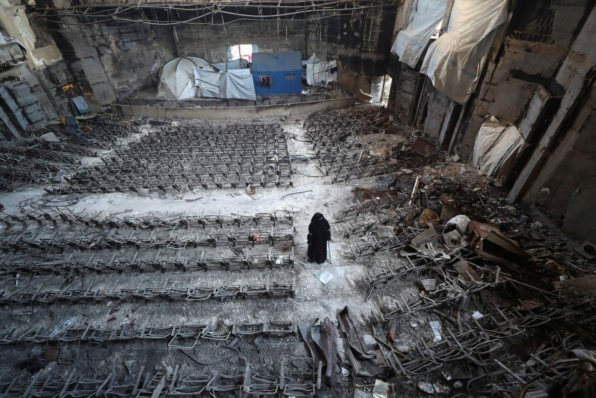 A woman in a black hijab stands on steps between rows of damaged seats in a lecture hall inside a ruined university building. 
