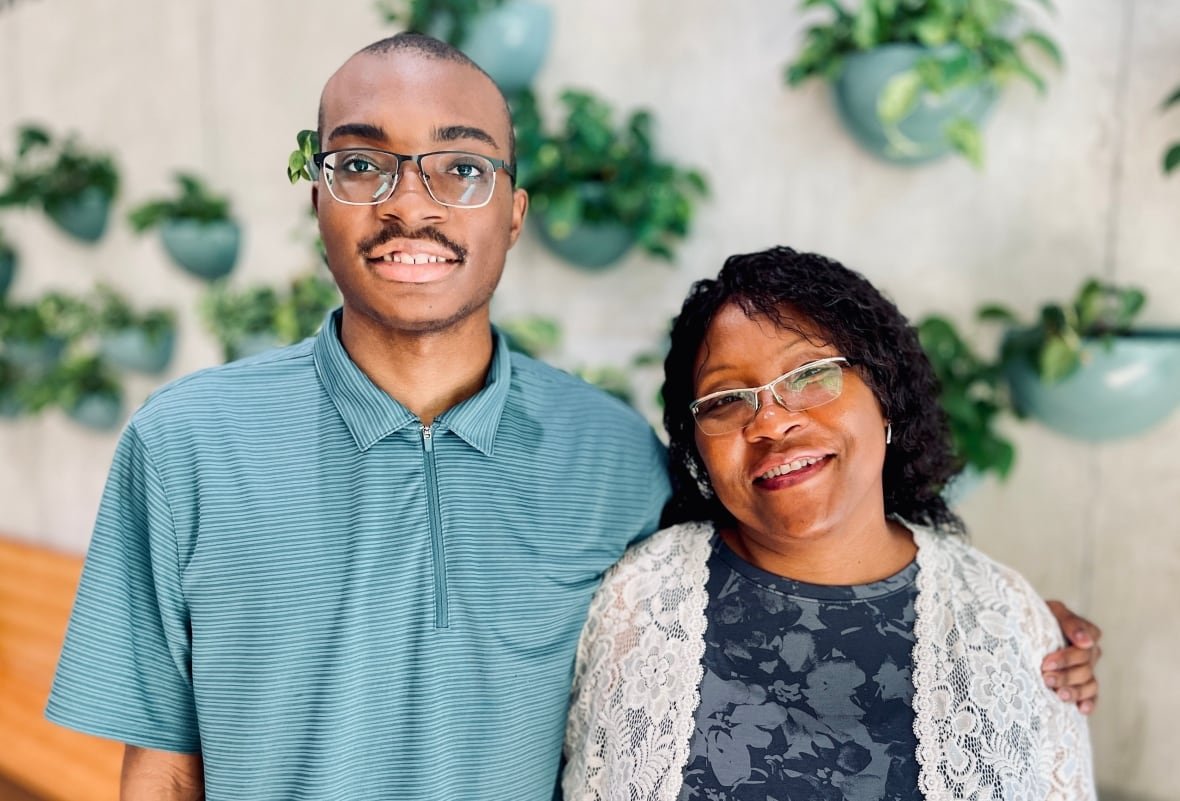 A smiling young man in glasses and a teal polo shirt stands with one arm around a smiling shorter woman wearing a blue floral-patterned dress with a white lace jacket. 