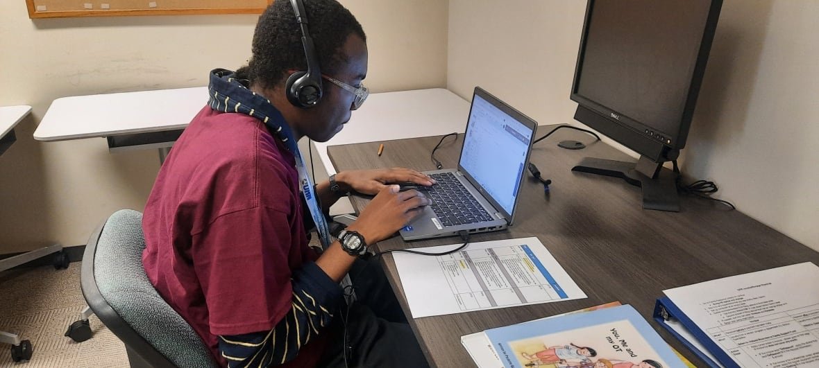 A young man in a burgundy tee-shirt with striped hoodie underneath peers at and types on a laptop computer while working a desk. 