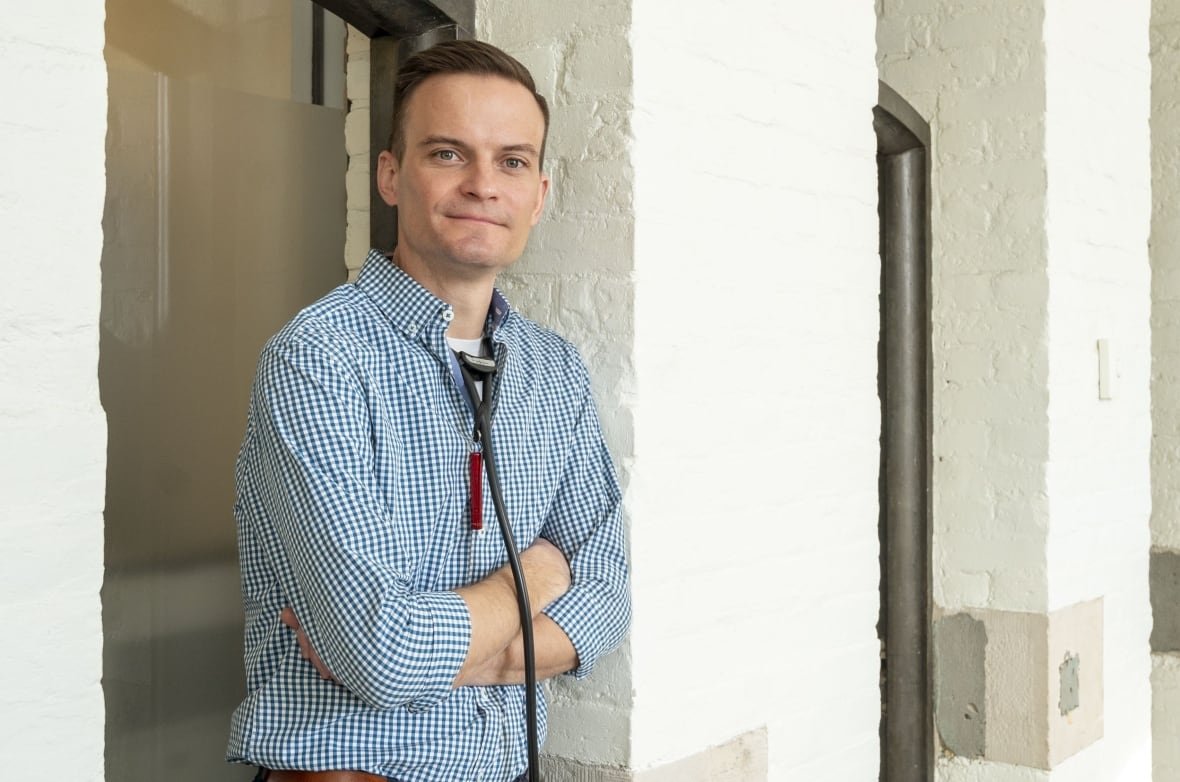 A man in a blue and white checkered dress shirt smiles with his arms folded.