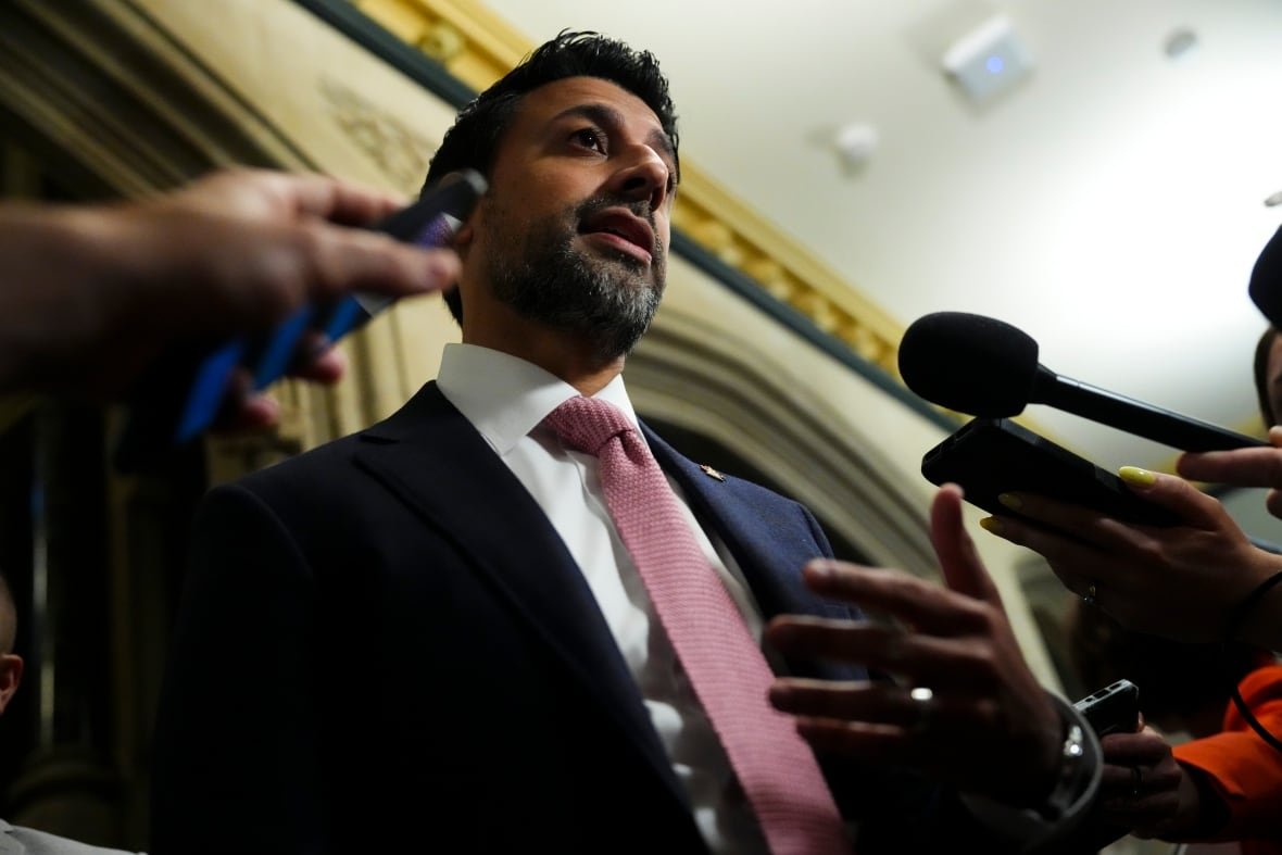 A man with dark skin and short black hair speaks into microphone. He's wearing a blue suit and pink tie