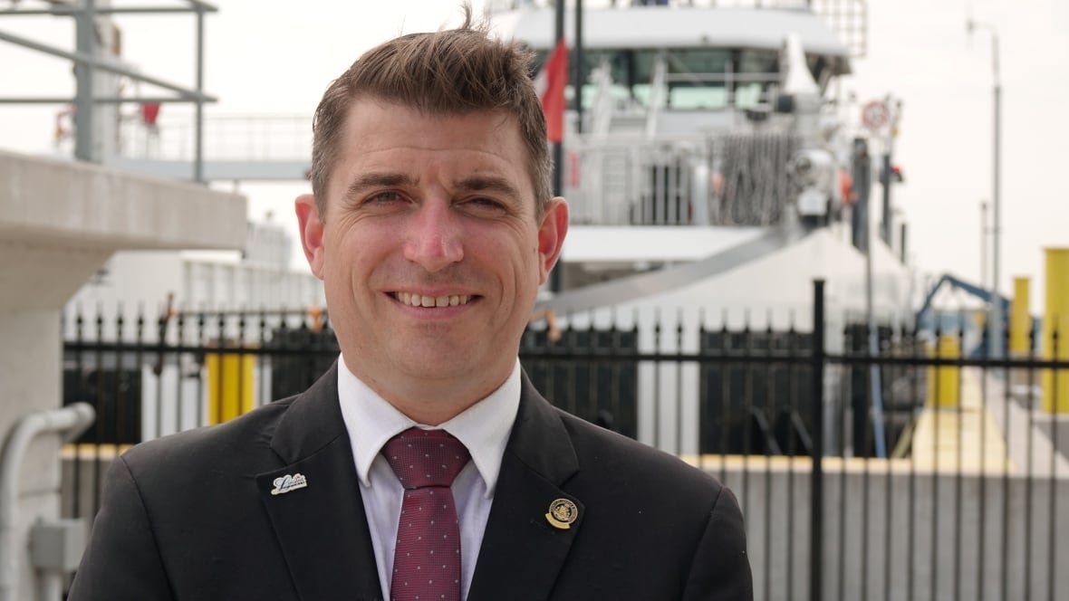 A man wearing a suit and tie smiles while standing in front of a large, white ferry boat.