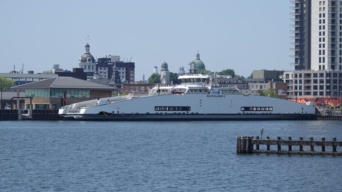 A large white ferry boat is shown on a sunny day. It's at a dock and behind it is a city skyline with high-rises and domed buildings.