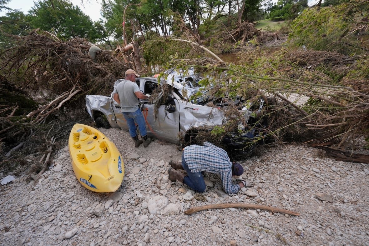 A man kneels to look under a destroyed truck that has a tree lying across its hood, while another man looks into the truck window