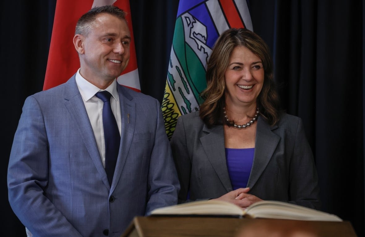 A white woman with brown hair is wearing a suit. She is smiling, while standing beside a smiling white man, with short grey hair, who is wearing a blue suit and tie.