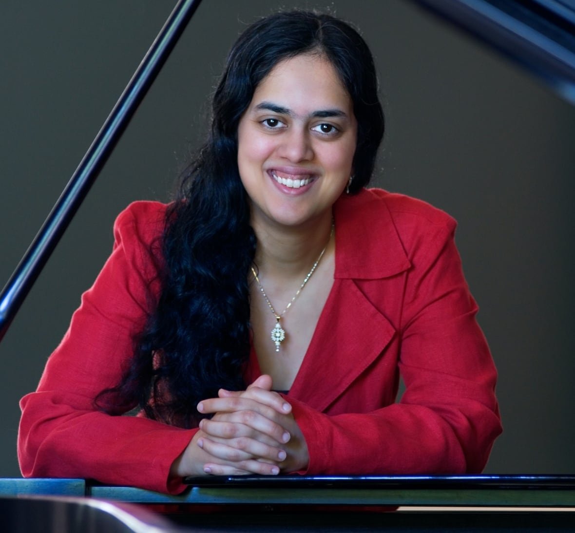 A woman with long black hair, wearing a red jacket, smiles as she sits at a grand piano.