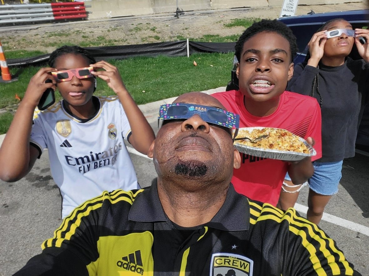 A man looks up wearing solar eclipse glasses. Behind him is a a young girl, wearing the same type of glasses, a boy holding a plate of food, and another young girl looking up wearing the solar ecliopse glasses.