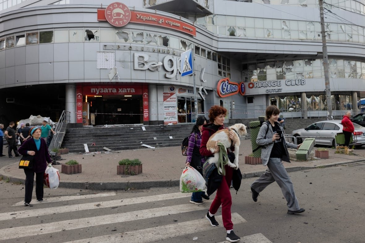 People emerge from a building in a city that shows signs of damage. They are carrying bags and various possessions. One woman is carrying a dog.