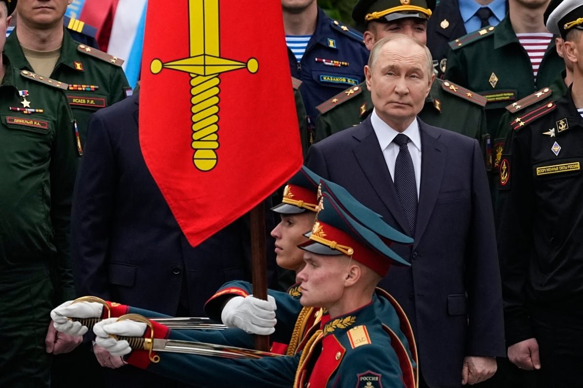 A man in a suit watches as men in military uniforms walk past. A large flag or banner to his right shows a golden sword on a rad background. 