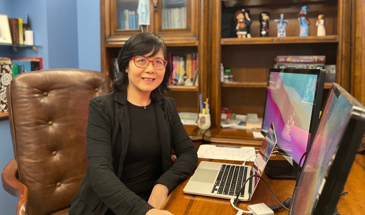 A woman in a black suit with read glasses, sits in front of a laptop in an office. Bookeshelves can be seen in the background.