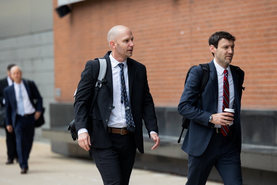Two people in suit and tie carrying bags are shown walking on a sidewalk outside a building.