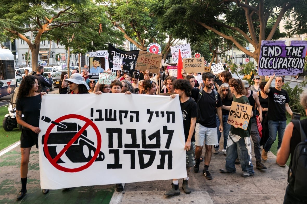 A few dozen persons are shown in an outdoor photo in an apparent protest, with banners in Hebrew shown.