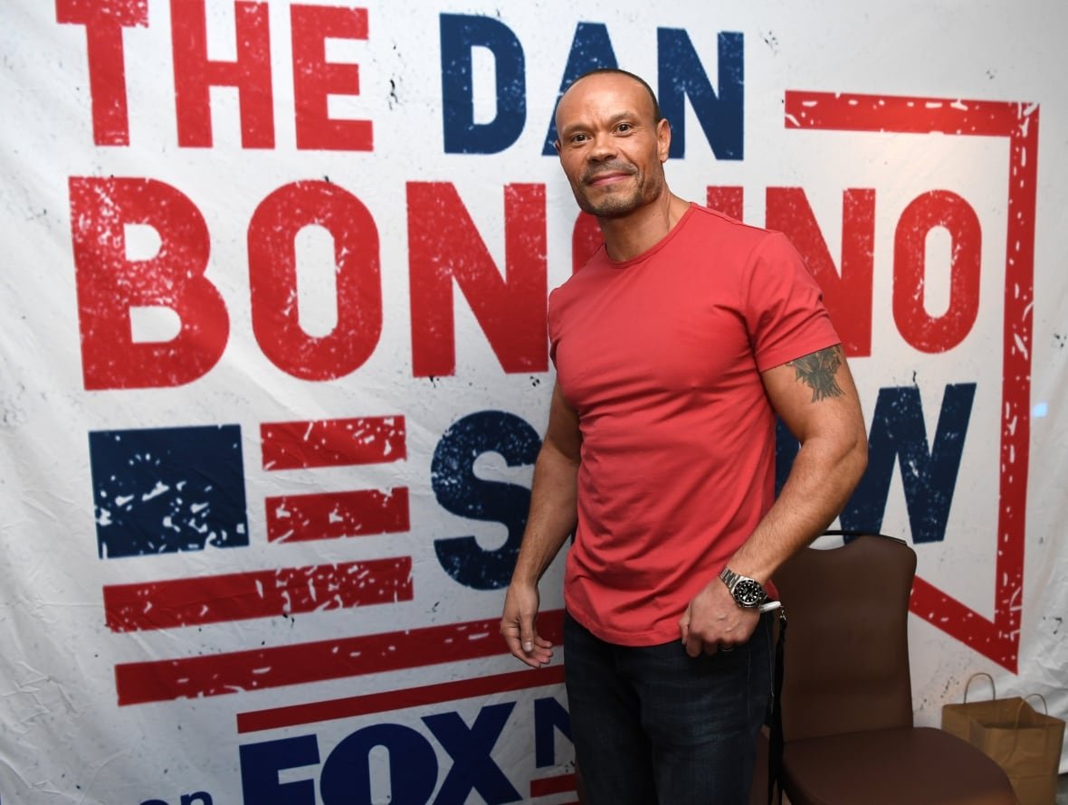 A balding man with a goattee wearing a tight t-shirt is shown standing before a backdrop that says The Dan Bongino Show.