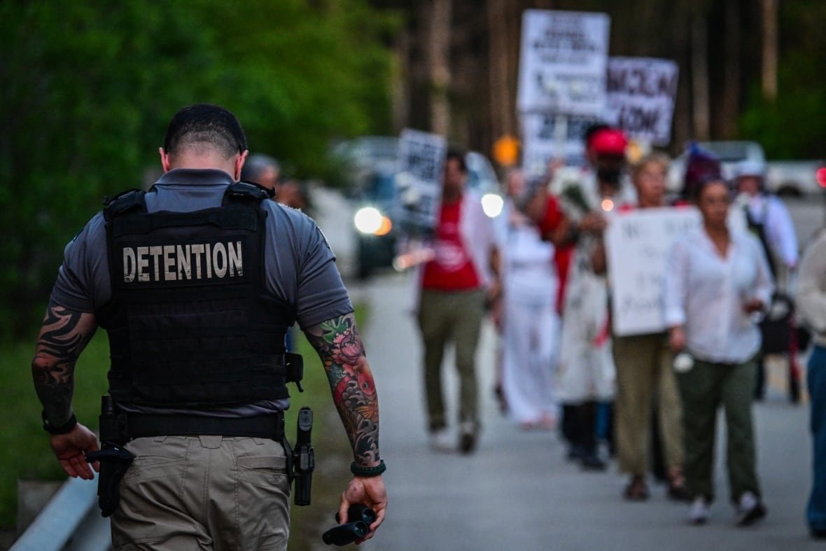 A tattooed, muscular man with a gun in a holsters is pictured from behind wearing a vest that reads "DETENTION" and walking toward a a group of protesters.