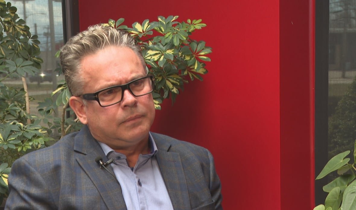 Man wears collared shirt and suit jacket as he sits near plants and a red wall. 