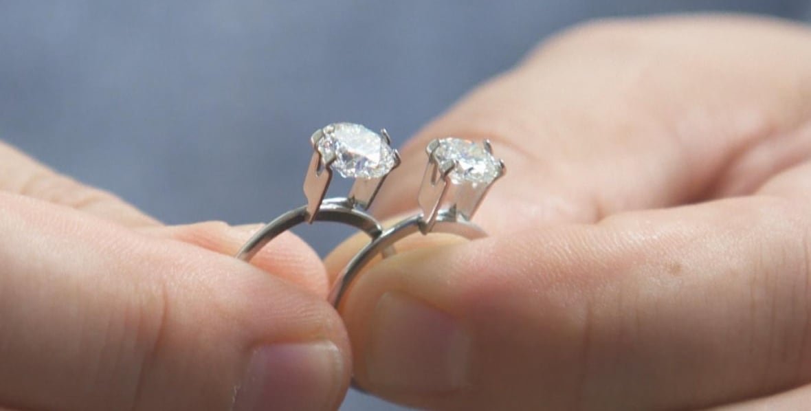 two diamonds in rings are shown close up, held in a person's two hands.