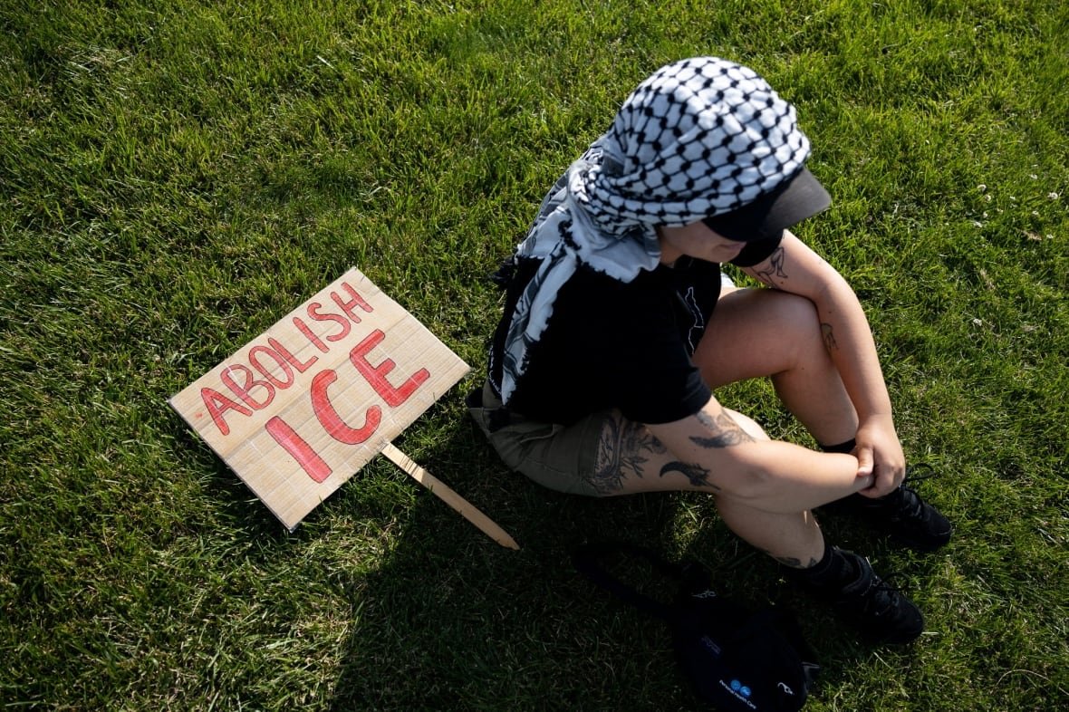 Individual sitting on the grass, head down, with a sign on the ground that says Abolish Ice.