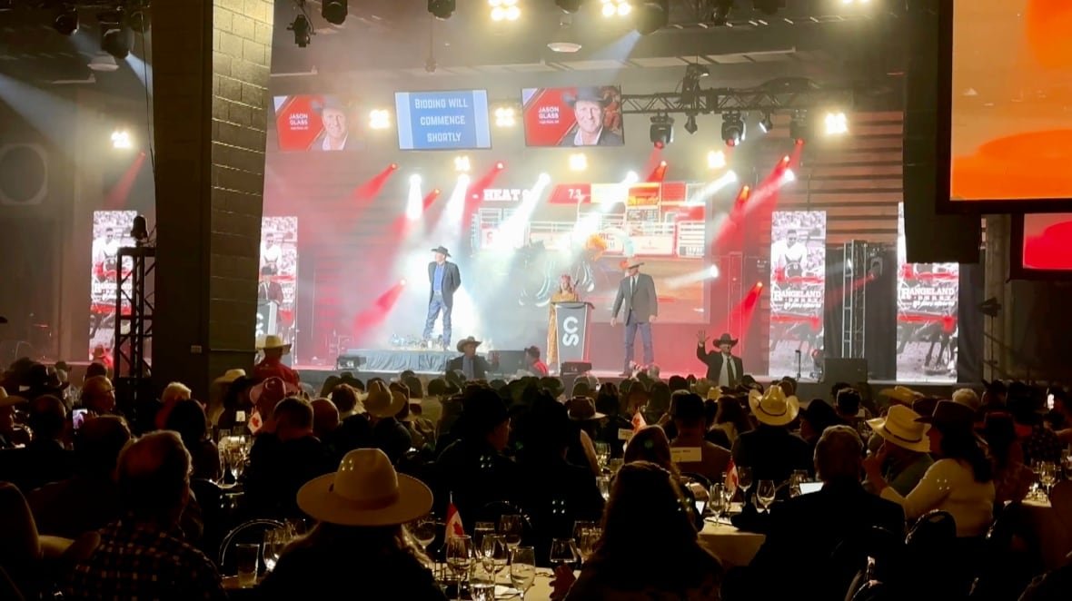 Bright white and red lights shine down on a stage as a man with a cowboy hat stands in front of a crowd.