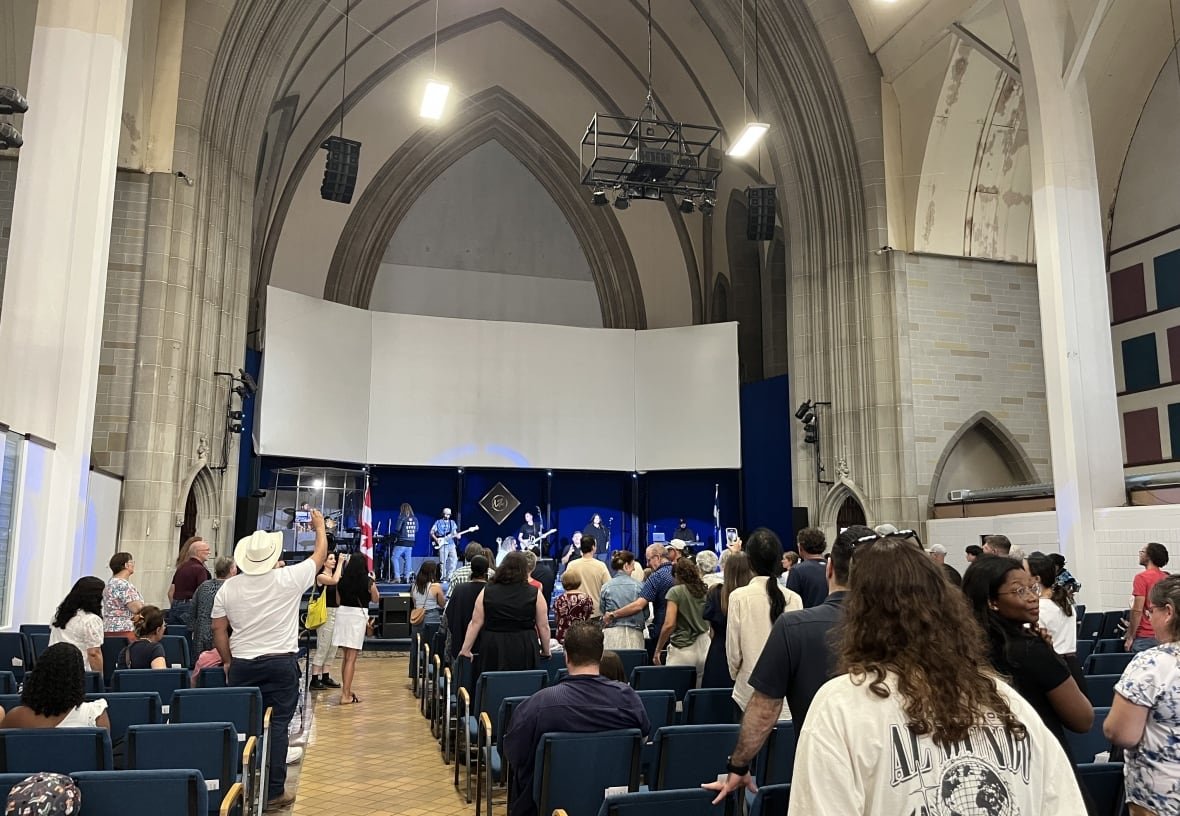 Musicians appear on a stage inside a church with worshippers sitting or standing as they look on.